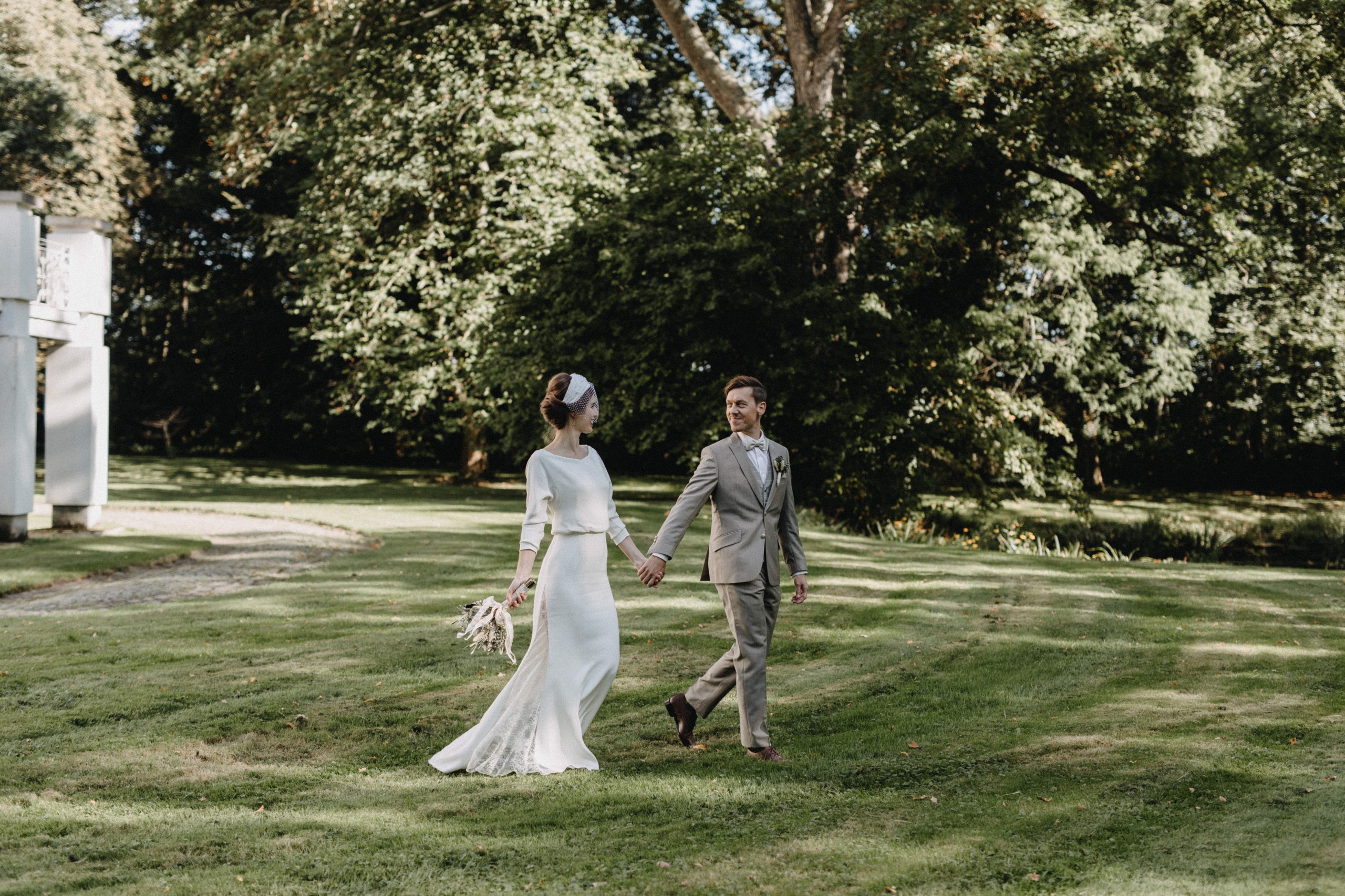 Braut und Bräutigam spazieren Hand in Hand über eine Wiese, umgeben von grünen Bäumen. Die Braut trägt ein elegantes, schlichtes Kleid und hält einen kleinen Blumenstrauß in der Hand. Beschriftung: Real Bride Anouk. Foto von Vanessa Sander.