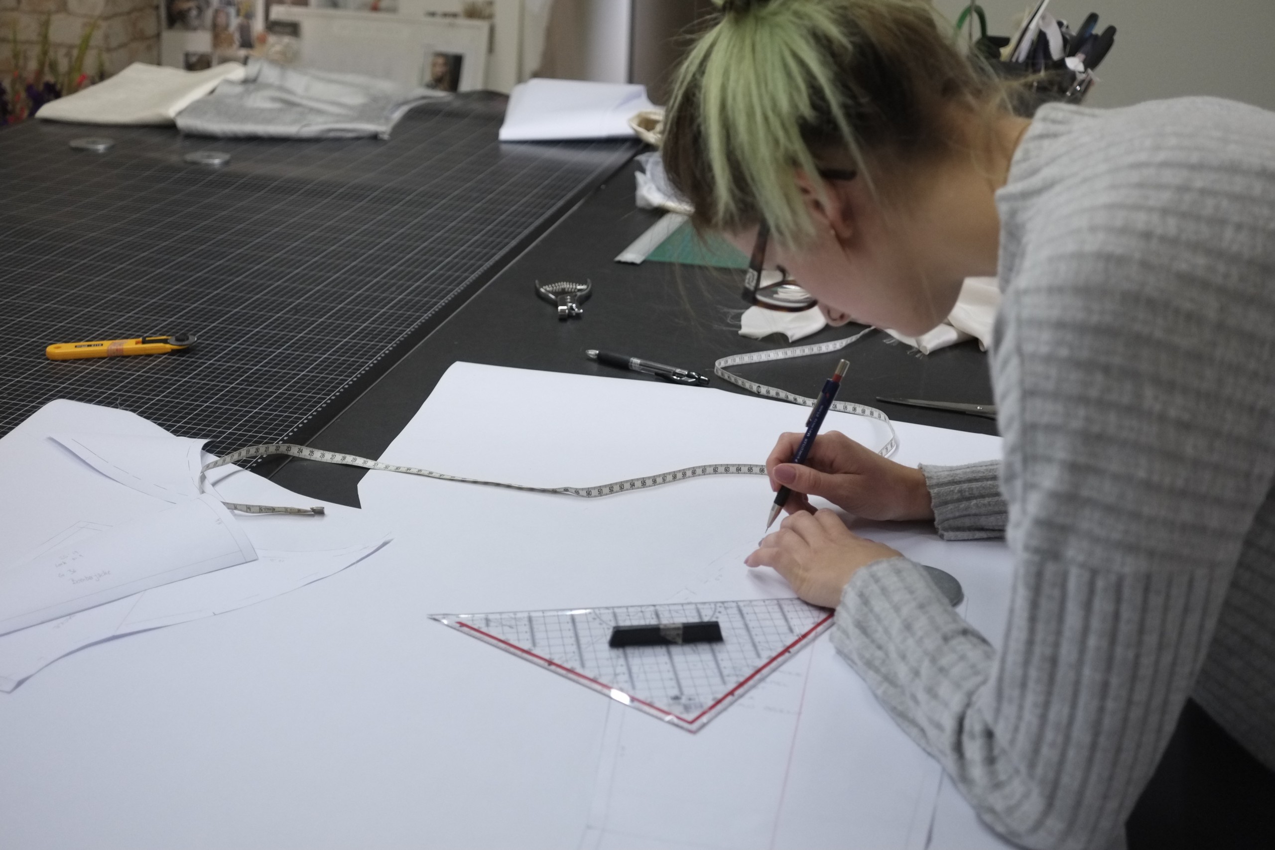 Person beim Zeichnen von Schnittmustern auf einem Tisch im Atelier, umgeben von Schneidewerkzeugen und Materialien. Foto aufgenommen im Atelier von Magdalena Mayrock, 2019.