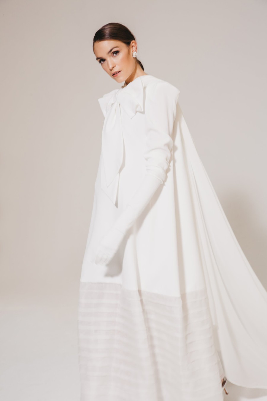 Braut in einem eleganten Brautkleid mit langen Ärmeln und Schleifendetail, modern und minimalistisch, mit fließendem Stoff und Cape-ähnlichem Design.