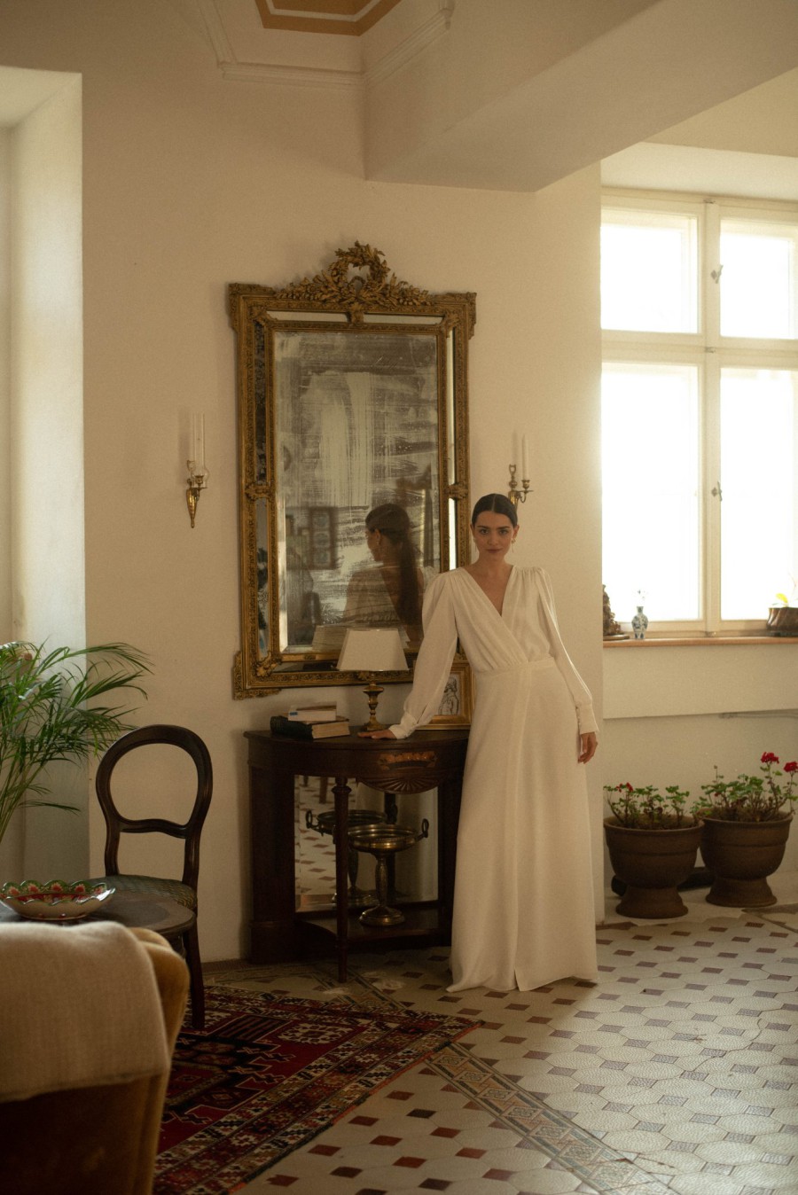 Braut in einem langen, fließenden Brautkleid, posiert in einem eleganten Raum vor einem antiken Spiegel.