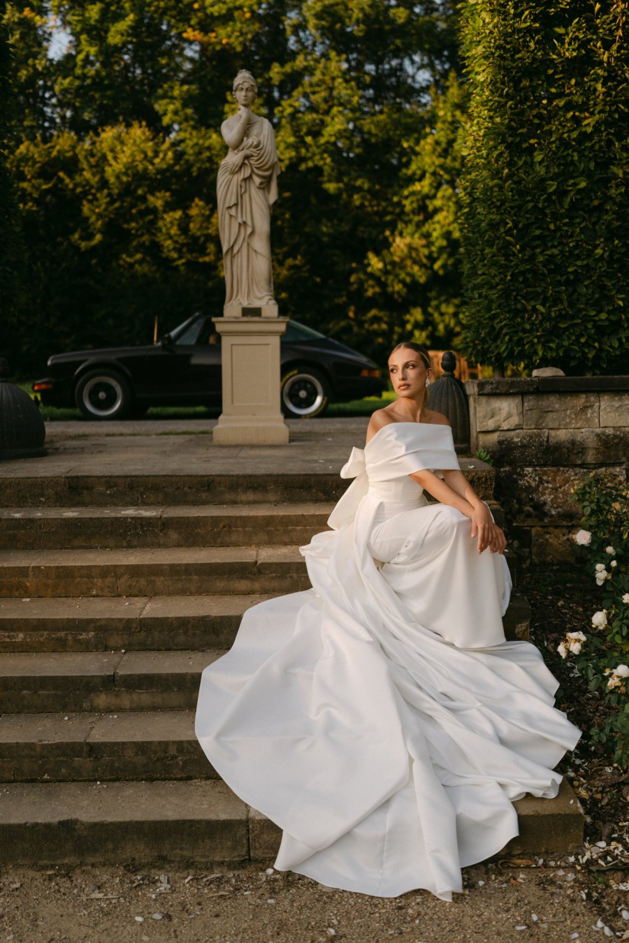 Braut in einem voluminösen, trägerlosen Brautkleid, sitzt auf einer Steintreppe in einem historischen Garten mit einer Statue im Hintergrund.