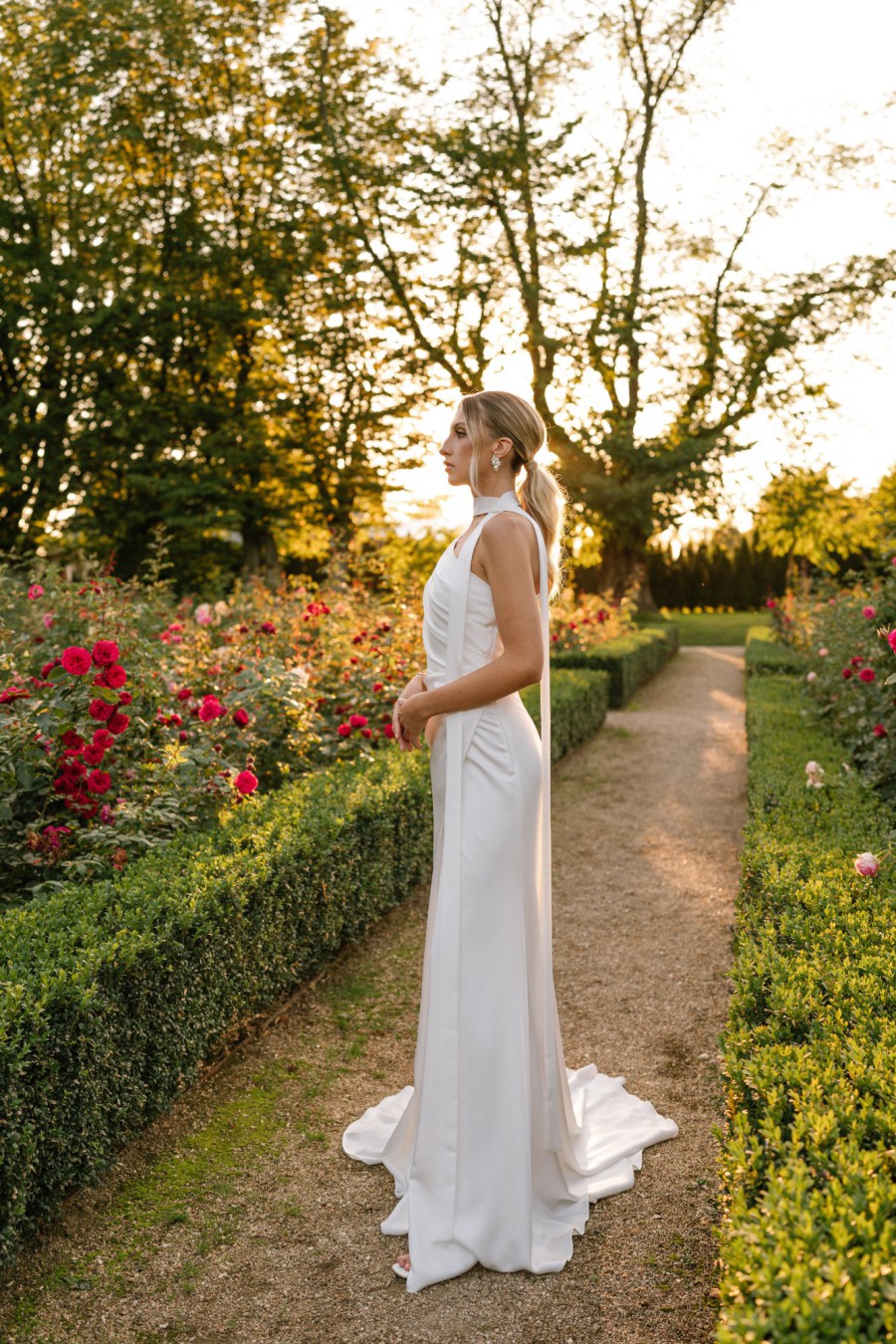 Braut in einem eleganten, schmal geschnittenen Brautkleid, posiert in einem blühenden Garten bei Sonnenuntergang.