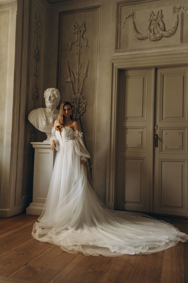 Braut in einem trägerlosen, bodenlangen Brautkleid mit langer Schleppe, posiert in einem eleganten, historischen Raum neben einer Statue.