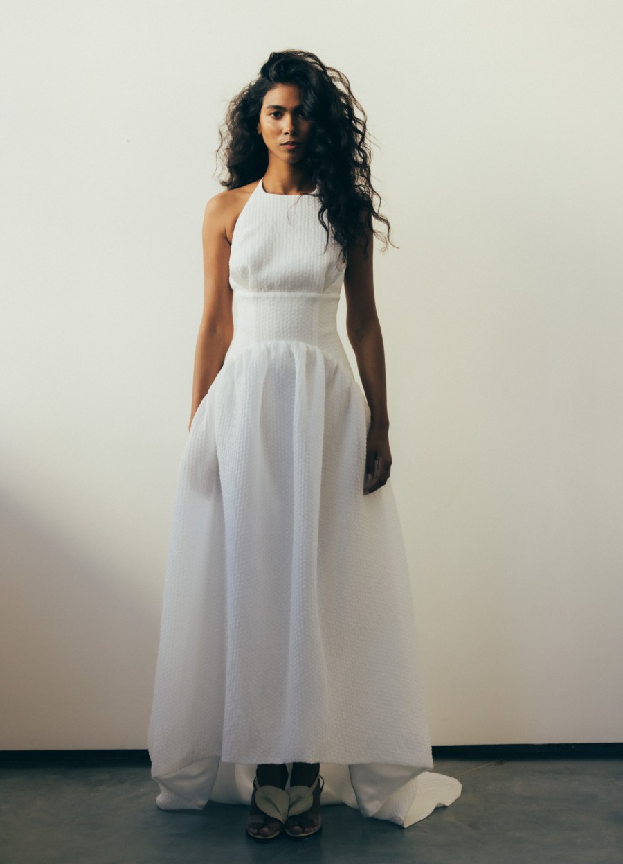 Modernes Neckholder-Brautkleid mit fließendem Rock, getragen von einer Frau mit lockigem Haar in einem minimalistischen Raum.