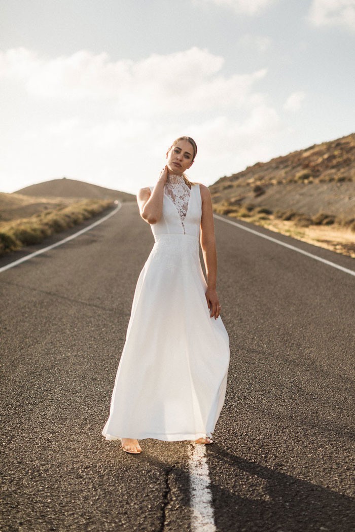 Eine Braut in einem langen, weißen Hochzeitskleid steht auf einer verlassenen Straße in einer offenen Landschaft unter hellem Himmel. Das Kleid hat einen hohen Ausschnitt mit zarten Spitzen-Details, während die Braut entspannt lächelt.