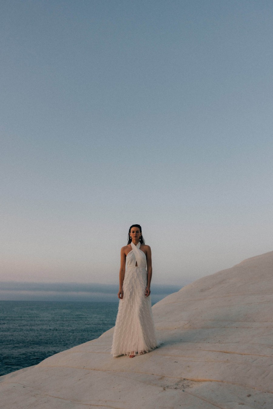 Braut in einem eleganten Neckholder-Brautkleid von Karolina Twardowska, steht auf einer Klippe mit Blick auf das Meer bei Sonnenuntergang.