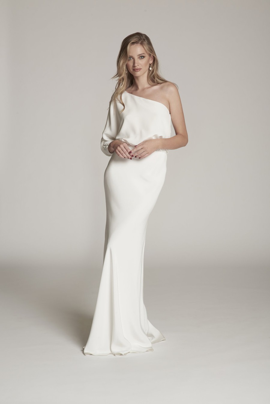 Braut in einem eleganten, einseitigen One-Shoulder-Brautkleid mit langem Ärmel, posiert vor einem neutralen Hintergrund.