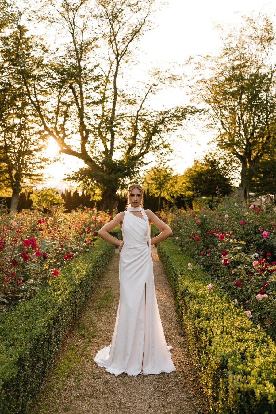 Braut in einem One-Shoulder-Brautkleid mit eleganter Schleppe, posiert in einem gepflegten Garten bei Sonnenuntergang.