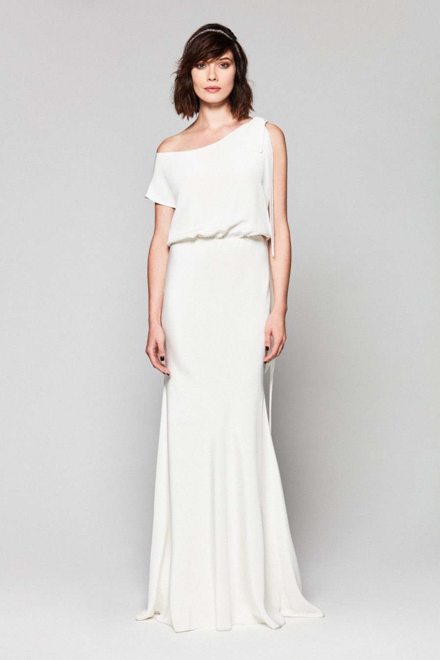 Braut in einem minimalistischen, One-Shoulder-Brautkleid mit schmaler Silhouette, posiert vor einem neutralen Hintergrund.