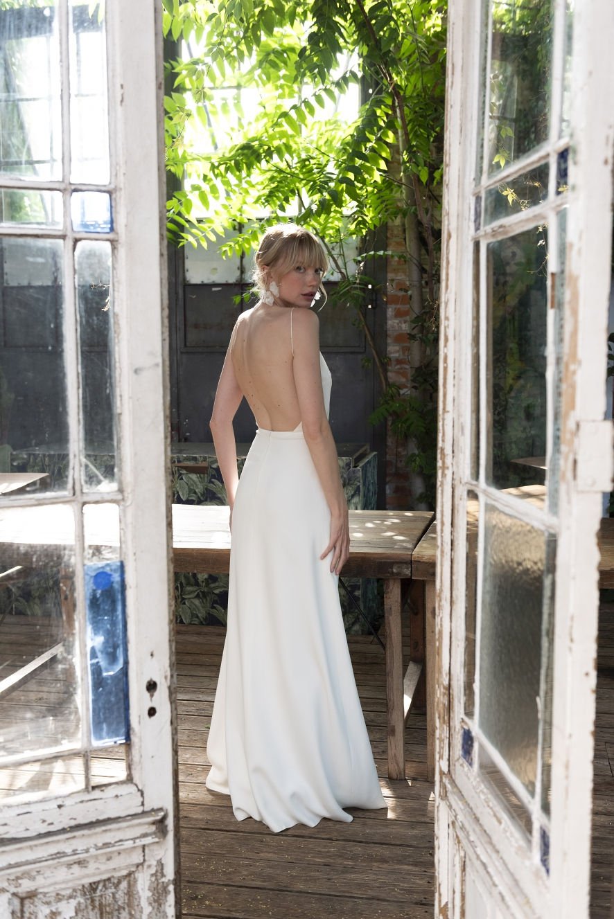 Farbfoto einer Frau in einem schlichten, rückenfreien Brautkleid, die durch eine offene Tür auf eine Terrasse tritt. Das Kleid hat eine schmale, bodenlange Silhouette und betont den offenen Rücken. Die Braut blickt über die Schulter zurück, während sie von Sonnenlicht durch die Bäume umgeben ist.