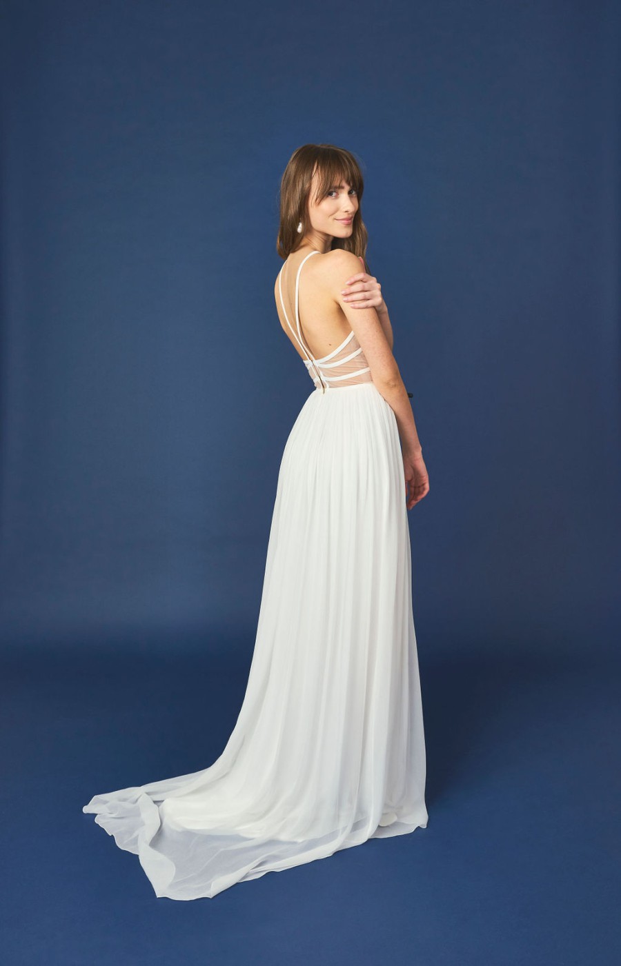 Frau in einem bodenlangen, rückenfreien Chiffon-Hochzeitskleid mit schmalen Trägern steht vor einem blauen Hintergrund. Das Kleid fällt sanft auf den Boden und bildet eine elegante Schleppe, die Bewegung in die Komposition bringt. Die Szene strahlt Romantik und Eleganz aus.