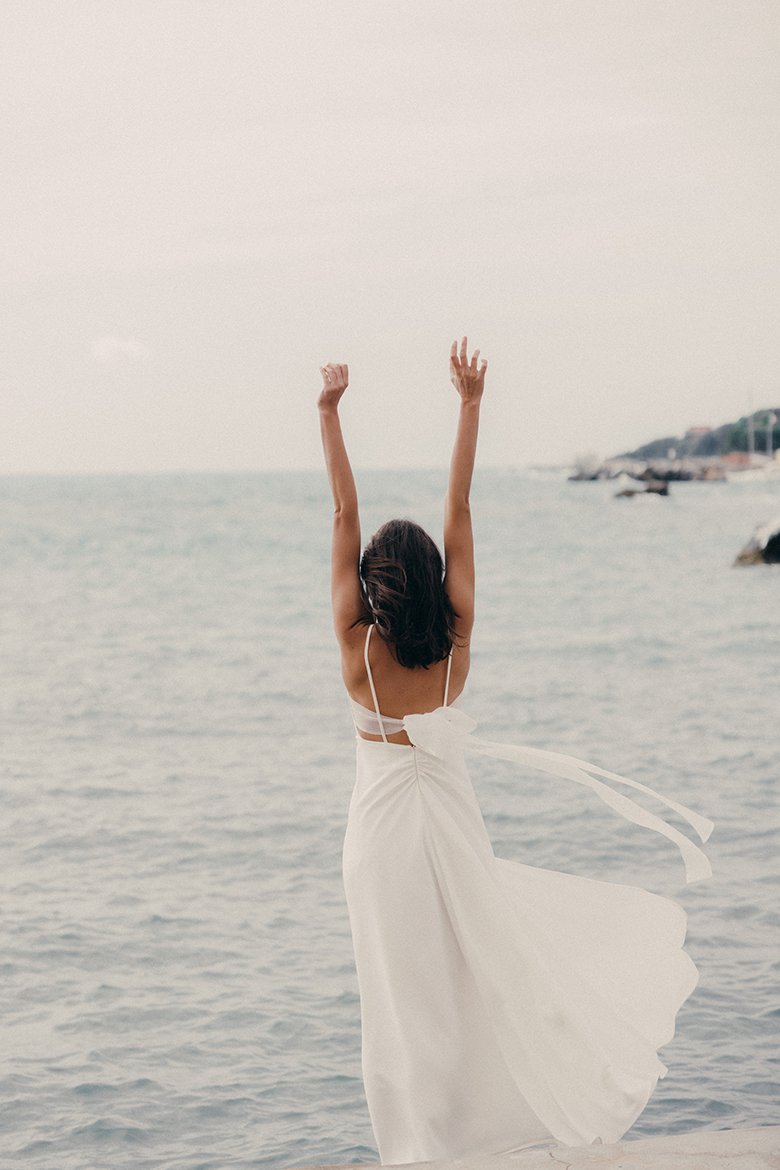 Frau in einem fließenden, rückenfreien Hochzeitskleid steht mit erhobenen Armen am Meer, während der Wind sanft den leichten Stoff ihres Kleides bewegt. Die Szene vermittelt ein Gefühl von Freiheit und Unbeschwertheit, mit einem malerischen, ruhigen Ozean im Hintergrund.