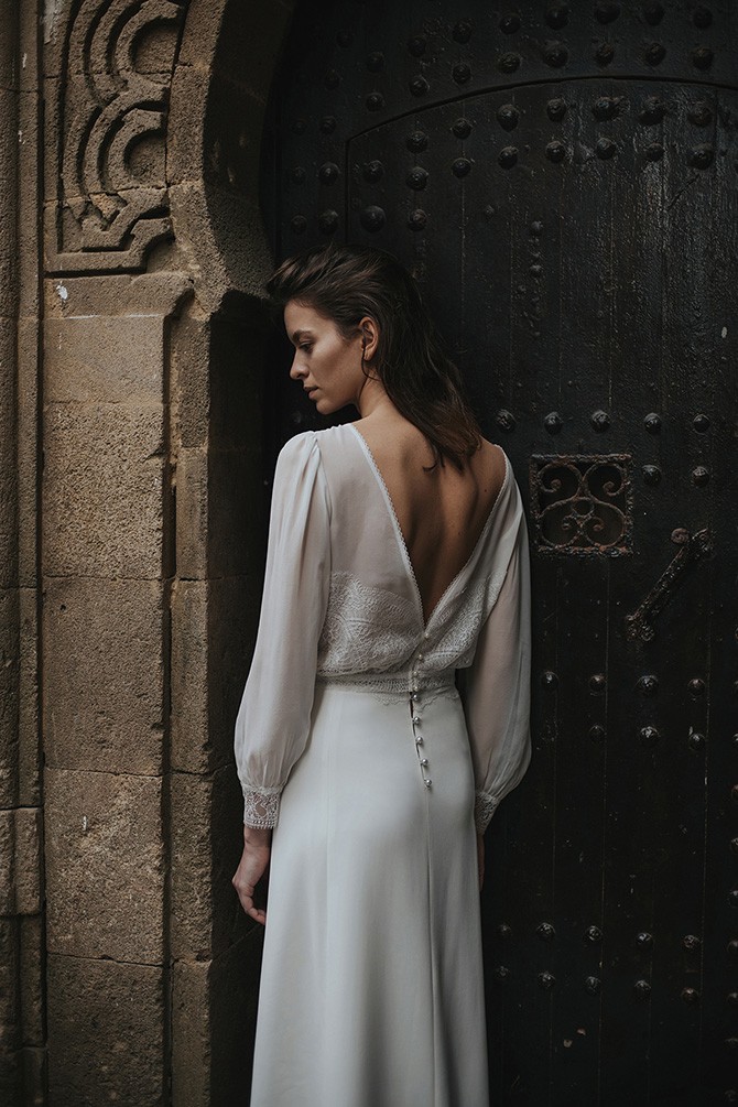 Frau in einem rückenfreien, romantischen Brautkleid mit langen Ärmeln, steht vor einem dekorativen, historischen Steintor. Das Kleid fällt in eleganten Falten nach unten und harmoniert mit der klassischen Architektur im Hintergrund, was eine Mischung aus Nostalgie und zeitloser Eleganz vermittelt.