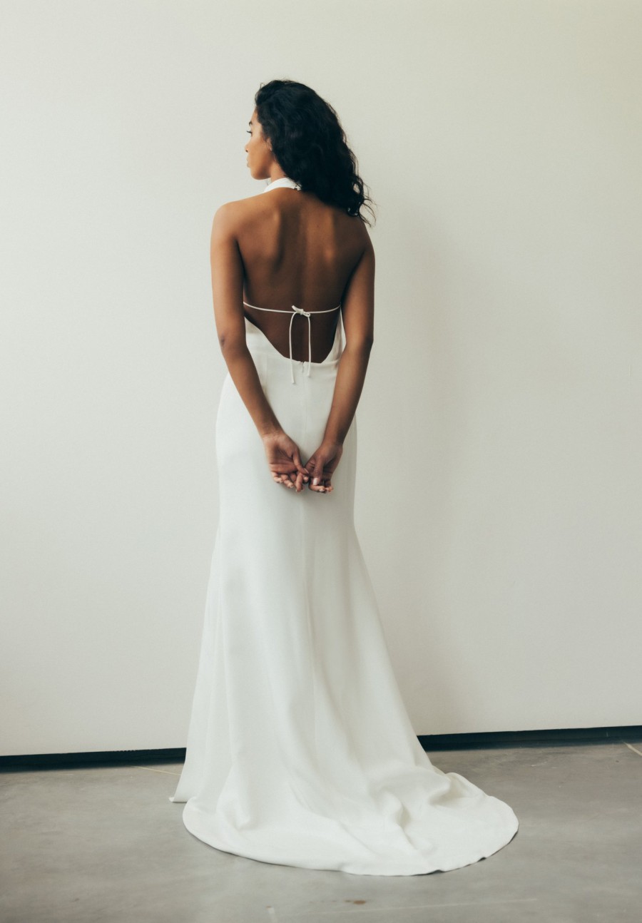 Farbfoto einer Frau in einem rückenfreien Brautkleid mit tiefem V-Rückenausschnitt. Das bodenlange Kleid hat eine schlichte, elegante Silhouette. Die Braut steht mit dem Rücken zur Kamera und hat ihre Hände locker hinter dem Rücken gefaltet, während ihr Haar über die Schulter fällt.