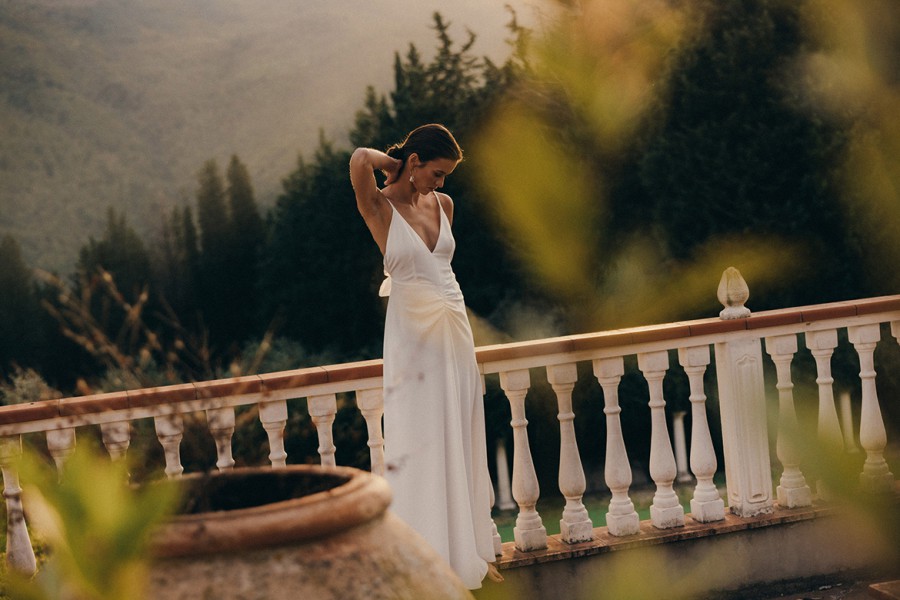 Eine Frau in einem schlichten, eleganten Brautkleid steht auf einer Veranda, die Hände in den Haaren, in Farbe fotografiert.