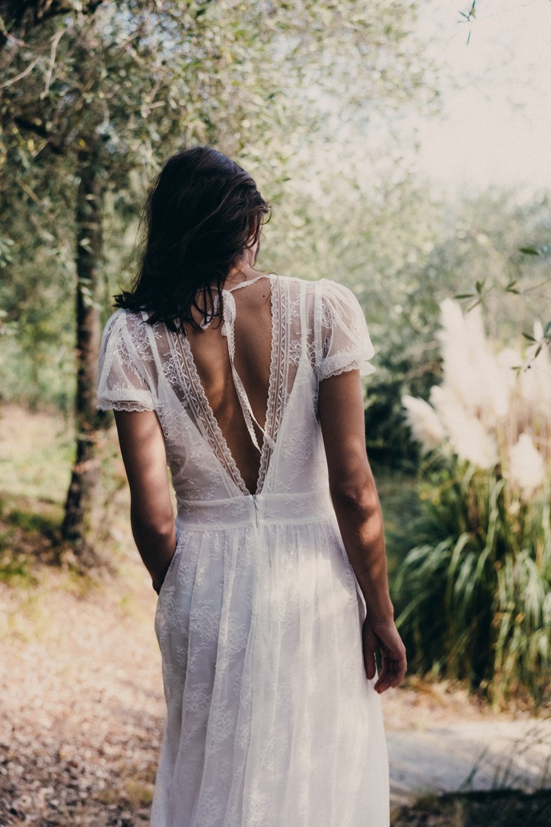 Eine Frau in einem Brautkleid aus Spitze mit tiefem Rückenausschnitt, durch einen Wald spazierend, in Farbe fotografiert.